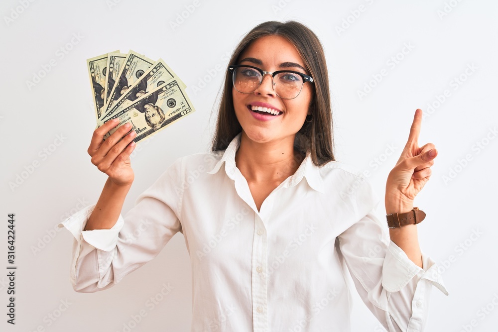 Beautiful businesswoman wearing glasses holding dollars over isolated white background very happy pointing with hand and finger to the side Beautiful businesswoman wearing glasses holding dollars over isolated white background very happy pointing with hand and finger to the side