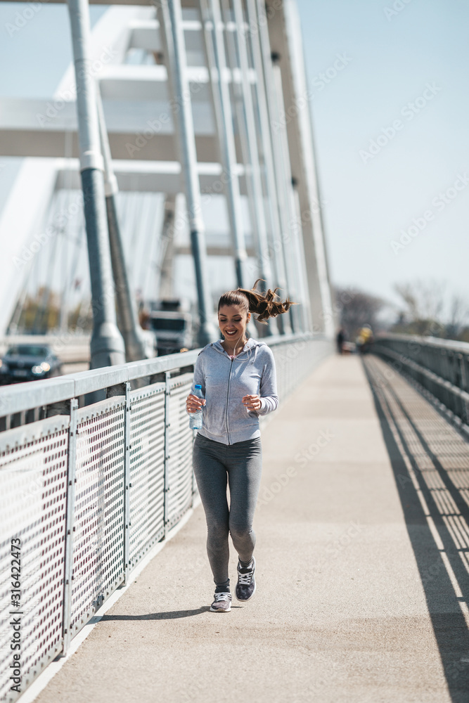 Beautiful young and fit woman in good shape running and jogging alone on city bridge street. She wears sporty earbuds and listens to music.