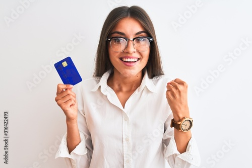 Young businesswoman wearing glasses holding credit card over isolated white background screaming proud and celebrating victory and success very excited, cheering emotion