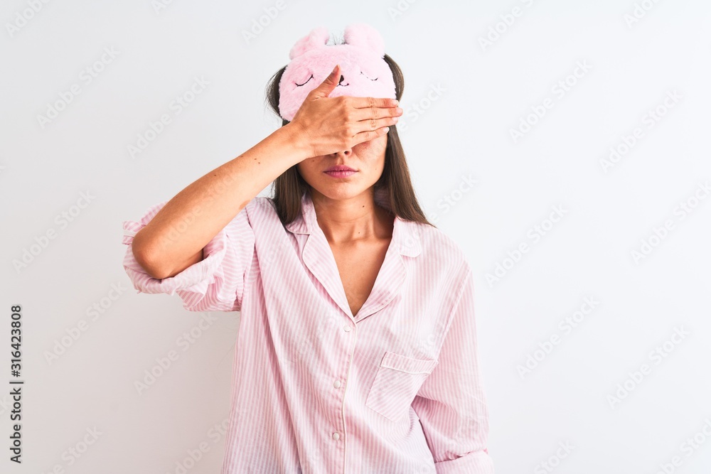 Young beautiful woman wearing sleep mask and pajama over isolated white background covering eyes with hand, looking serious and sad. Sightless, hiding and rejection concept Young beautiful woman wearing sleep mask and pajama over isolated white background covering eyes with hand, looking serious and sad. Sightless, hiding and rejection concept