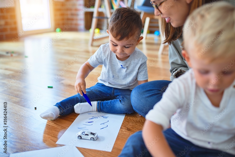 Beautiful teacher and group of toddlers sitting on the floor drawing using paper and pencil around lots of toys at kindergarten - obrazy, fototapety, plakaty 