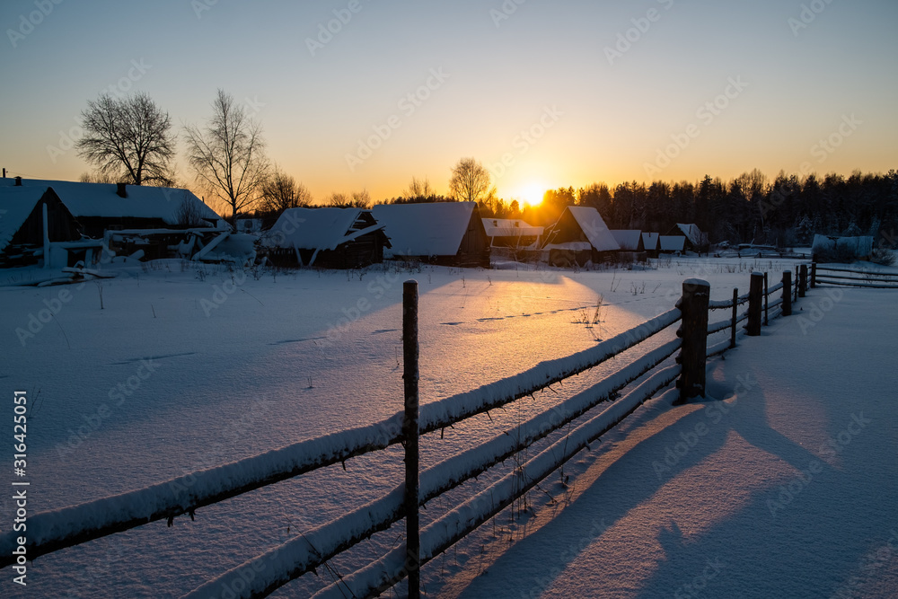 rural landscape. The sun sets behind the forest and creates contour shadows on the snow. The fence goes into the distance and creates a perspective