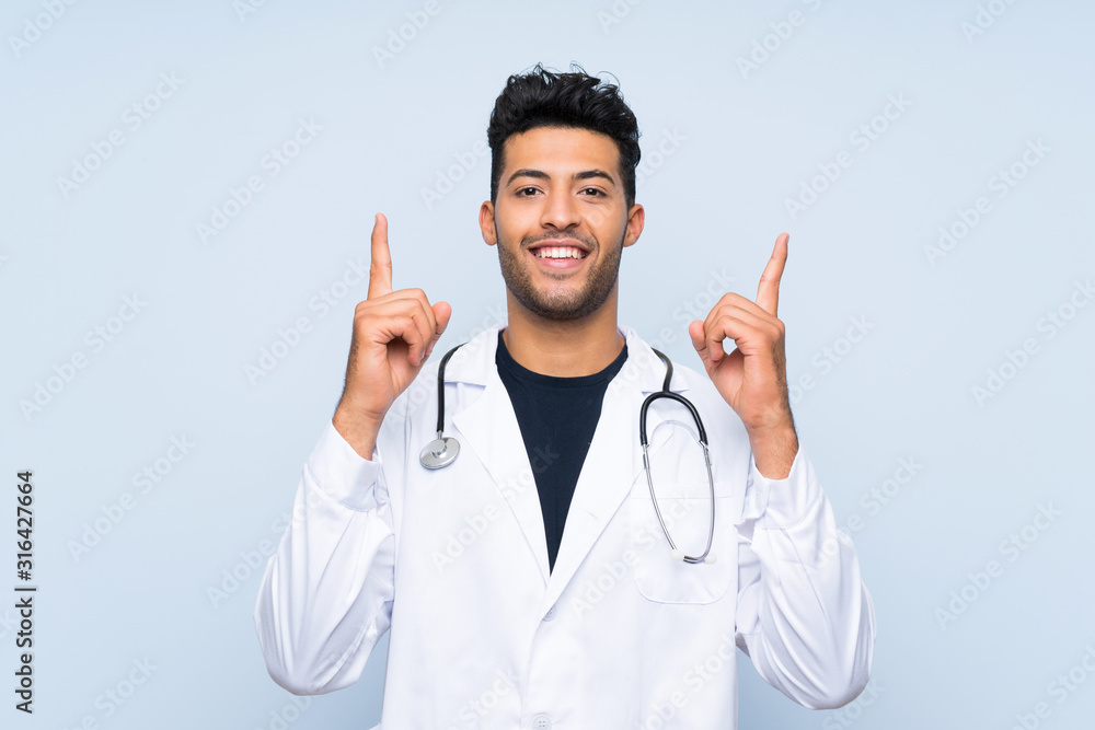 Young doctor man over isolated blue wall pointing up a great idea Young doctor man over isolated blue wall pointing up a great idea