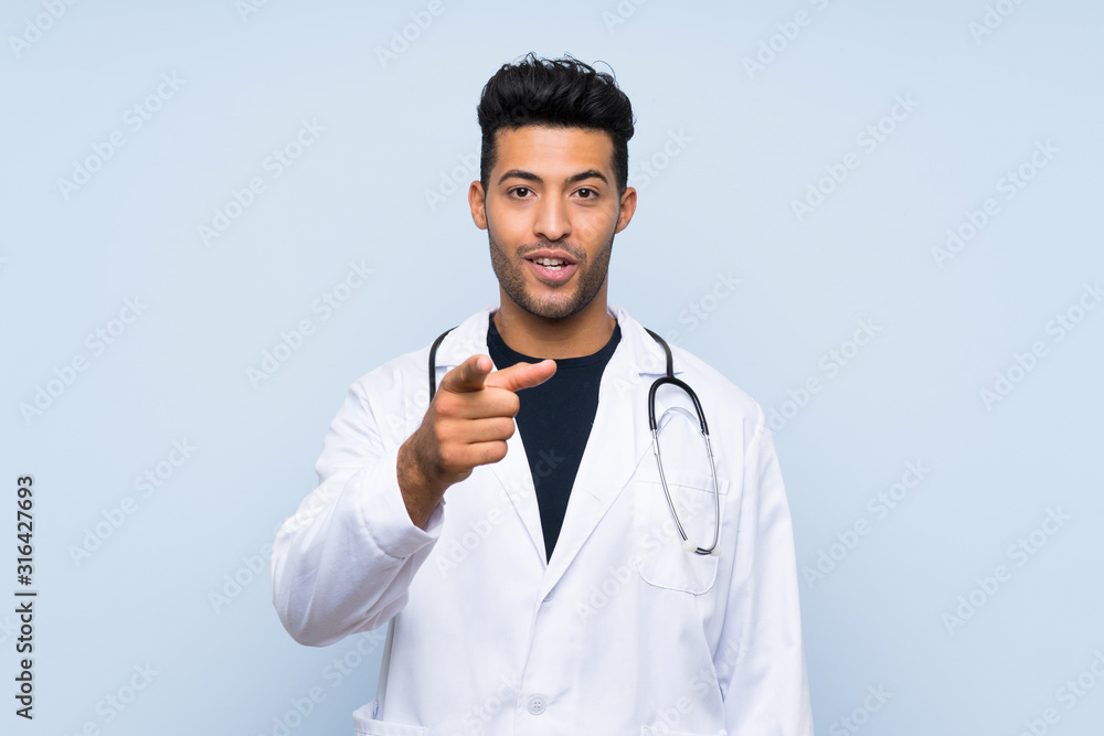 Young doctor man over isolated blue wall surprised and pointing front Young doctor man over isolated blue wall surprised and pointing front