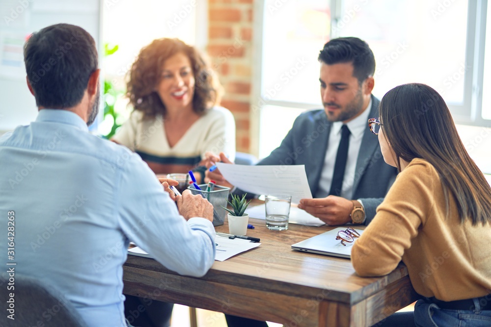 Group of business workers smiling happy working together. Sitting on desk with smile on face reading documents at the office Group of business workers smiling happy working together. Sitting on desk with smile on face reading documents at the office