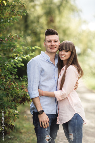 Young romantic couple is having fun in summer sunny day in park. Enjoying spending time together in holiday. Man and woman are hugging and smiling.