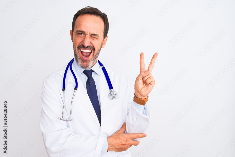 Middle age doctor man wearing coat and stethoscope standing over isolated white background smiling with happy face winking at the camera doing victory sign. Number two.