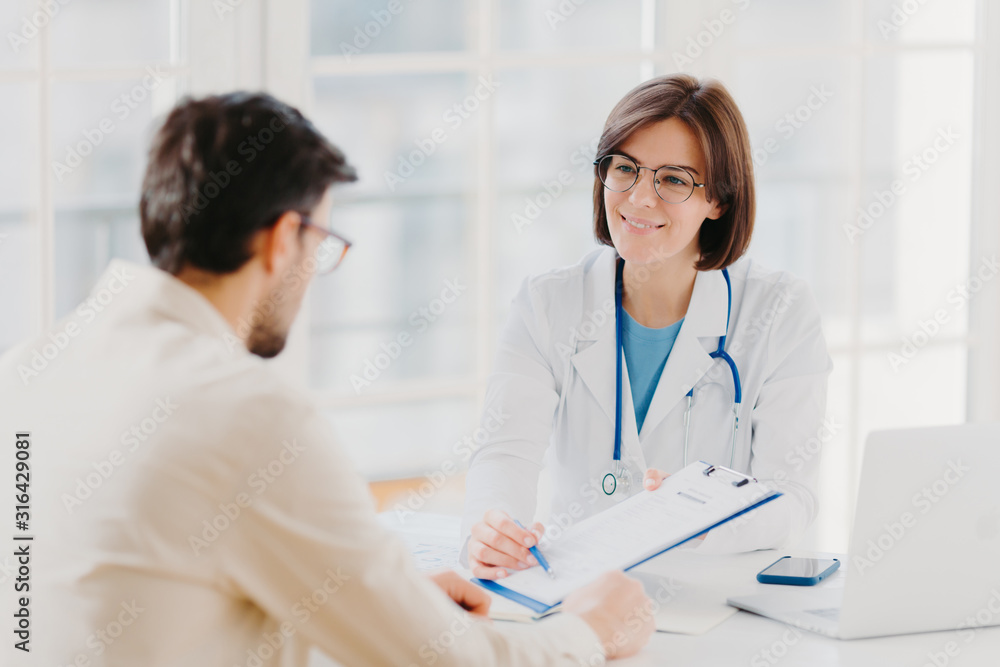 Young female doctor consults patient, shows information and prescription in clipboard, talk about medical checkup, pose at clinic, explains health insurance contract, read service agreement. Young female doctor consults patient, shows information and prescription in clipboard, talk about medical checkup, pose at clinic, explains health insurance contract, read service agreement.