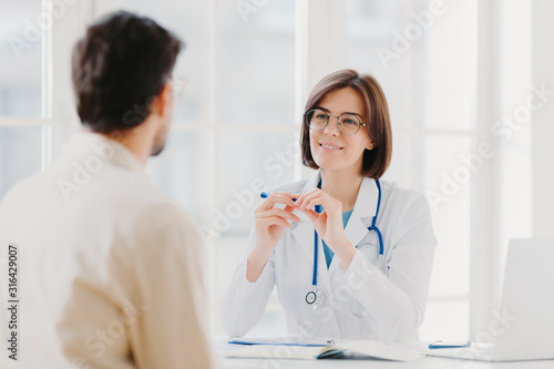 Professional female doctor in coat listens attentively patients symptoms  examines health  writes down information in patient card  pose in clinic. Sick male tells complaint  sits back to camera