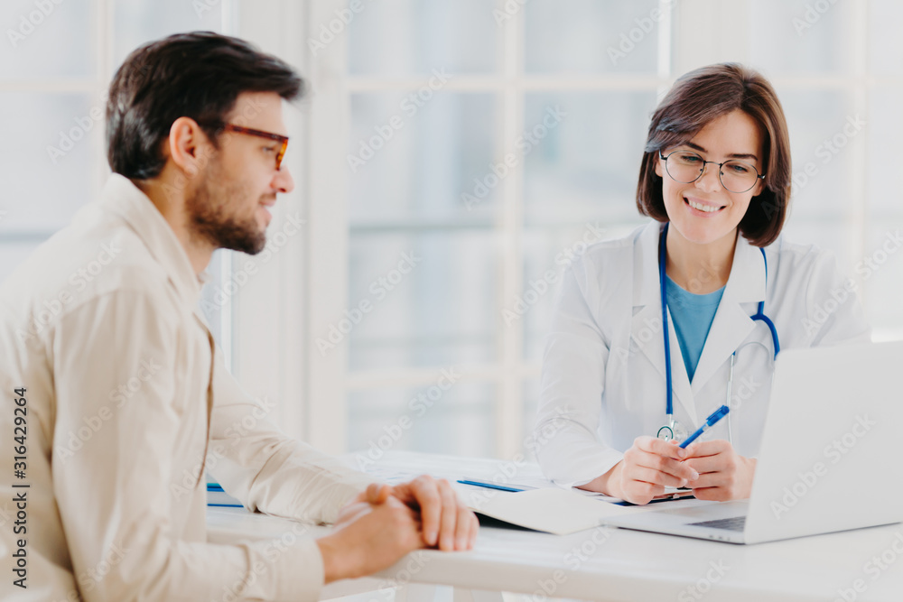 Horizontal shot of doctor and patient discuss something, talk about diagnosis, use laptop computer, pose in consulting room, check medical records. Sick man gets prescription from therapist. Horizontal shot of doctor and patient discuss something, talk about diagnosis, use laptop computer, pose in consulting room, check medical records. Sick man gets prescription from therapist.
