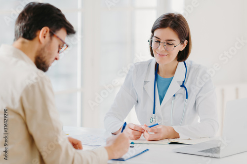 Male patient writes down application form, gets consultancy and advice about home treatment from doctor, use laptop and medical document at desk, pose at office together. Man doctors appointment