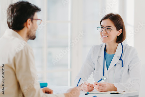 Photo of male patient writes all details makes notes during consultancy with doctor sit in spacious office. Physician tells treatment issues and explains diagnosis to hospital visitor. Medical concept