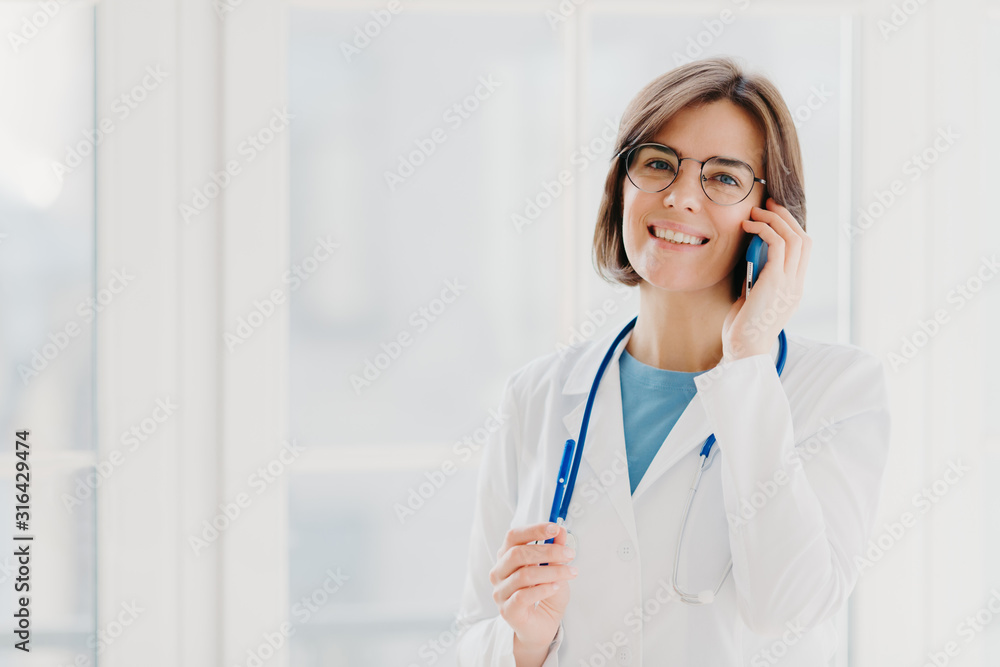 Positive woman doctor consults patient via cellphone, looks gladfully at camera, holds pen, wears white coat, poses in medical office, poses near window. Healthcare and consultation concept. Positive woman doctor consults patient via cellphone, looks gladfully at camera, holds pen, wears white coat, poses in medical office, poses near window. Healthcare and consultation concept.