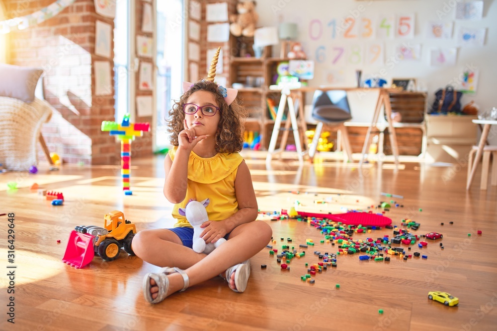 Beautiful toddler wearing glasses and unicorn diadem sitting on the floor at kindergarten with hand on chin thinking about question, pensive expression. Smiling with thoughtful face. Doubt concept.