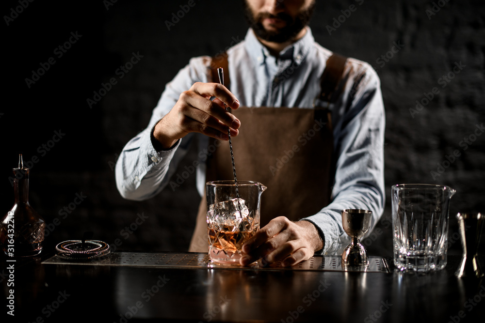 Male bartender stirring a brown cocktail with ice cubes in the glass with special spoon
