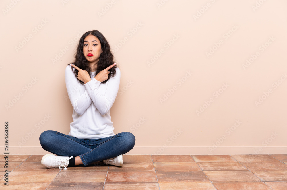 Young woman sitting on the floor pointing to the laterals having doubts Young woman sitting on the floor pointing to the laterals having doubts