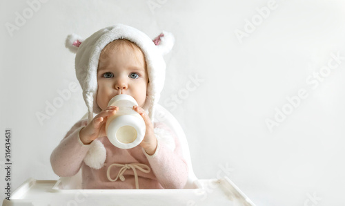 Little toddler girl in a warm fluffy hat drinks milk from a bottle while sitting. Half-length portrait. White gray background