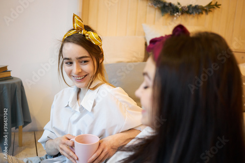 Pretty teenage girls chatting at home, sharing secrets and discussing plans for weekend. Happy young woman with headscarf sitting on floor in bedroom, having coffee and speaking to her female friend