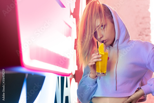 Nihgtlife. Teen girl standing isolated on club neon sign drinking cocktail posing to camera confident