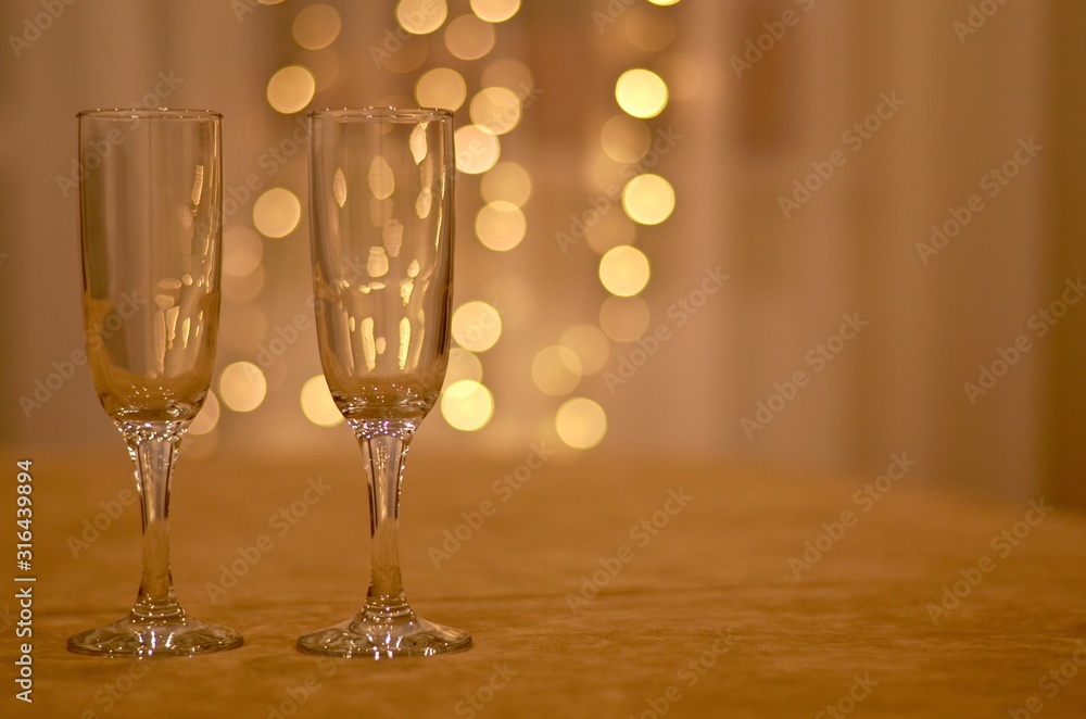 Two transparent wine glasses a soft beige surface with a dim light background of white curtains and lanterns garlands.