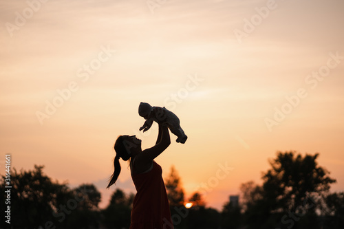 Sunset Silhouette: Young mother holding her baby boy child in city park standing in front of setting sun and vivid orange sky - Family values warm color summer scene handheld