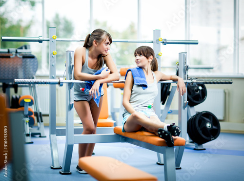 Two young beautiful slim women in grey sportswear with towels with bottle of water and looking at each other in gym. Healthy active lifestyle, sport, bodycare concept