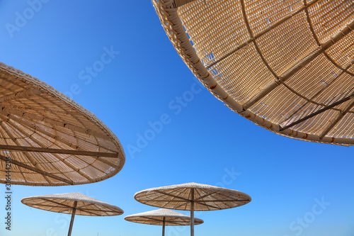 Fototapeta Naklejka Na Ścianę i Meble -  Tropical beach sun shades against bright blue sky on a sunny day. Up view. Sun umbrellas of reed and straw. Travel and vacation. Background with copy space
