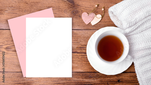 Mock up romantic composition with empty paper blank, heart decor, cup of tea, white knitted scarf on wooden background. Greeting concept for Valentines day, Mothers day or wedding card. Banner. 