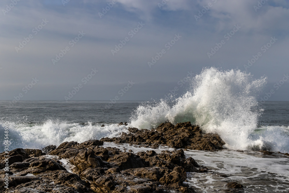 Wave breaking on rocky shoreline at Leo Carrillo State Beach, California. Rocks in foreground; pacific ocean, cloudy blue sky in background. 