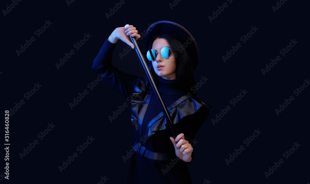 Portrait of mysterious sensual beautiful brunette woman in black futuristic dress, hat and glasses. High fashion model posing in studio on dark background. Blue neon light. Free space for text.
