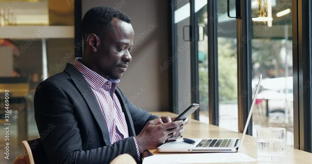 Young African Businessman typing on mobile phone with business clients in office laptop on the table by the window freelance Black man checking information on cellphone chatting on phone in cafe