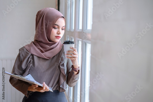 Beautiful modern Arabic business woman in brown hijab hand holding business documents and drinking coffee in office workplace.