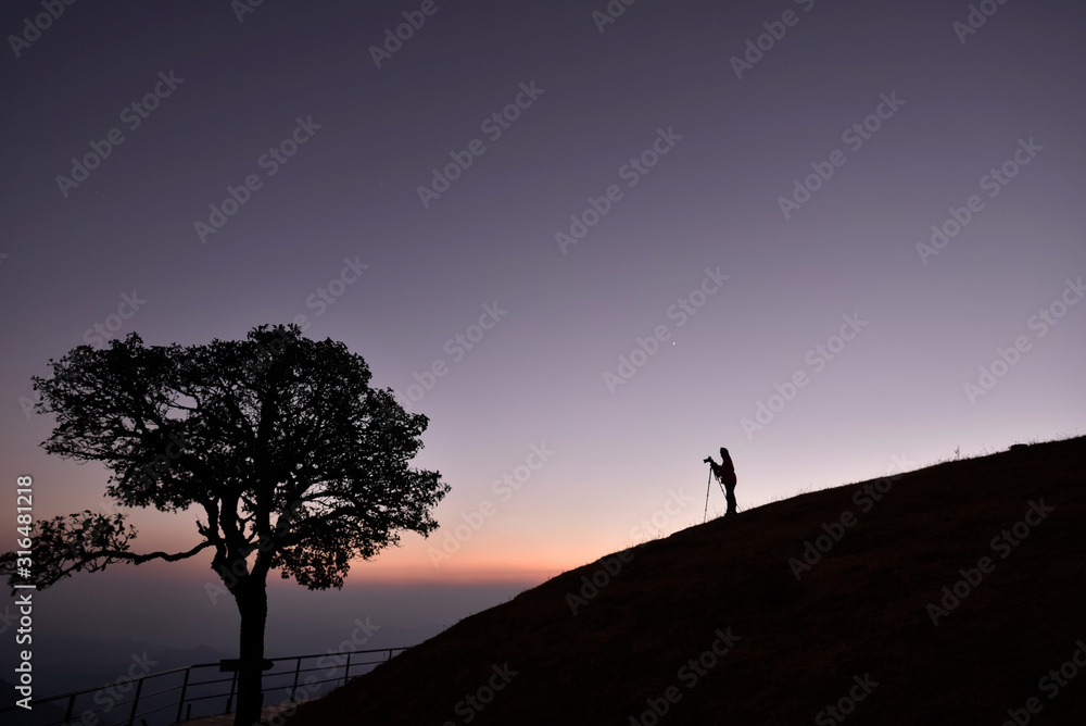 A photographer silhouette (with camera and tripod) on a hill with twilight sky background in an evening