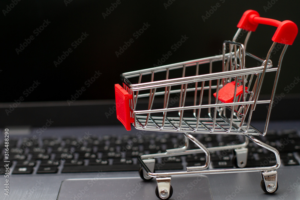 Small red shopping cart on silver laptop for shopping online with black screen background.