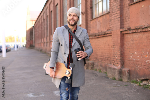 Handsome young man in grey coat and hat walking on the street, using longboard.