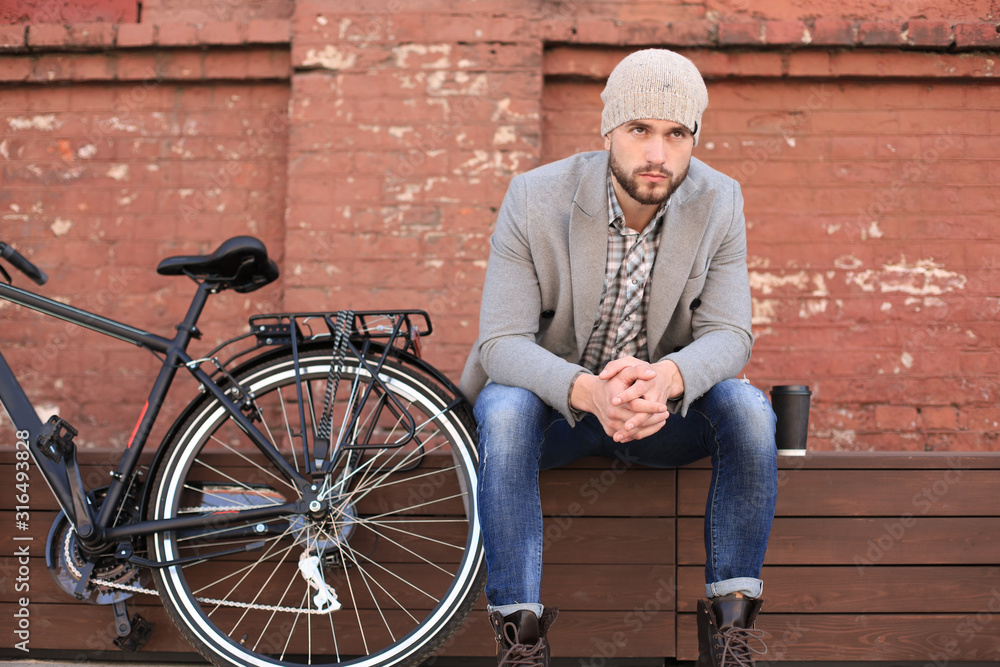 Handsome young man in grey coat and hat sitting on a bench relaxed drinking coffee and thinking near his bicycle. Handsome young man in grey coat and hat sitting on a bench relaxed drinking coffee and thinking near his bicycle.