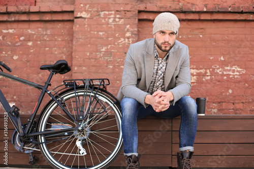 Wallpaper Mural Handsome young man in grey coat and hat sitting on a bench relaxed drinking coffee and thinking near his bicycle. Torontodigital.ca