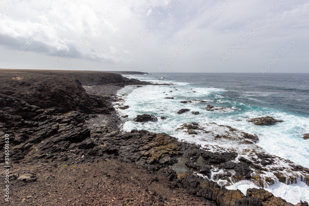 Scenic view at the coastline in the natural park of Jandia (Parque Natural De Jandina) on canary island Fuerteventura with gravel, lava rocks and rough sea with waves Scenic view at the coastline in the natural park of Jandia (Parque Natural De Jandina) on canary island Fuerteventura with gravel, lava rocks and rough sea with waves