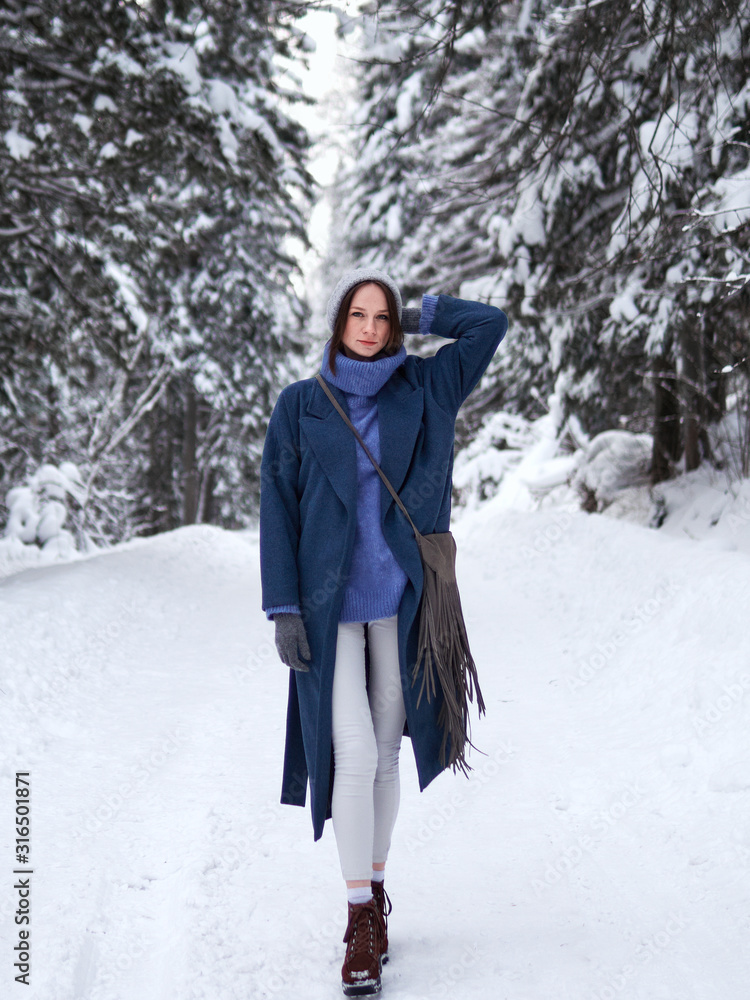 brunette walks through the winter forest in a gray hat, blue sweater and blue coat. brunette walks through the winter forest in a gray hat, blue sweater and blue coat.
