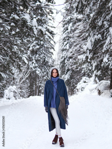 brunette walks through the winter forest in a gray hat, blue sweater and blue coat.