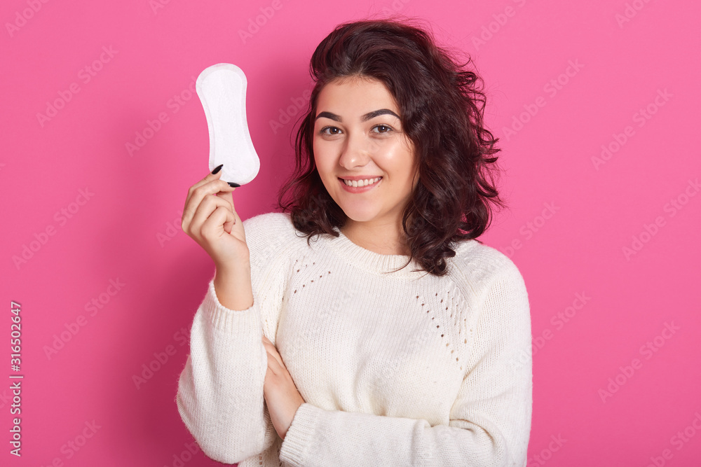 Positive delighted cute brunette looking directly at camera, smiling sincerely, having pleasant facial expression, holding hygiene pad in one hand, having menstruation. Women and health concept. Positive delighted cute brunette looking directly at camera, smiling sincerely, having pleasant facial expression, holding hygiene pad in one hand, having menstruation. Women and health concept.