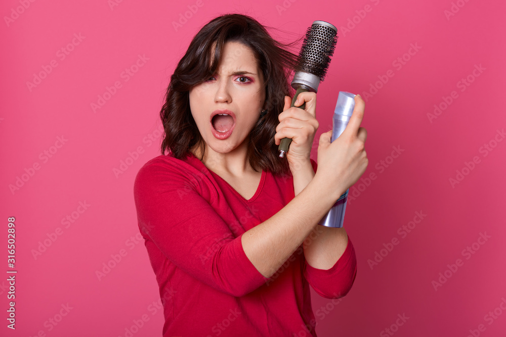 Close up portrait of young displeased emotional woman having indignation, opening eyes and mouth widely, looking directly at camera, dealing with hair with help of hairbrush and liquid for fixation. Close up portrait of young displeased emotional woman having indignation, opening eyes and mouth widely, looking directly at camera, dealing with hair with help of hairbrush and liquid for fixation.