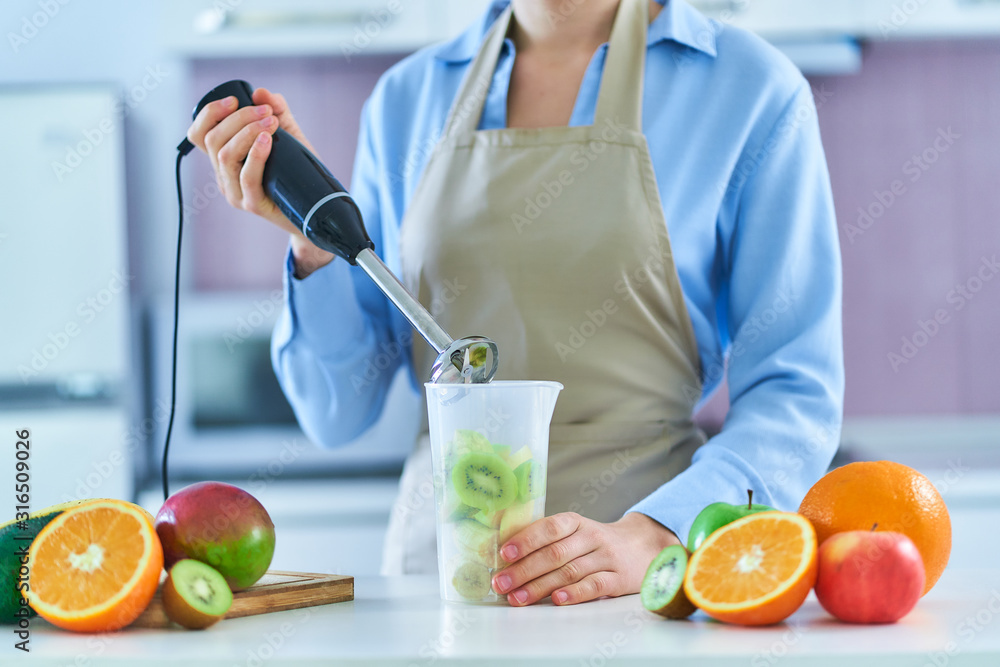 Female in apron using hand blender for preparing a fresh organic fruit smoothie at home in the kitchen