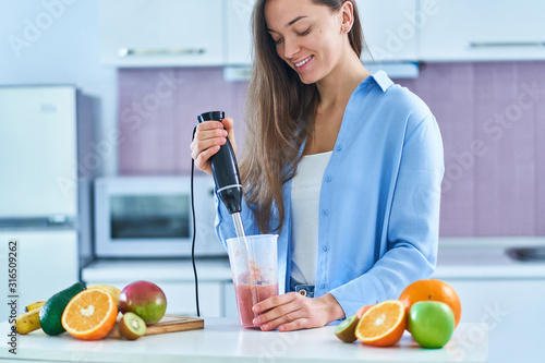 Photos Happy cute female uses hand blender to mixing fresh organic fruits for prepare d