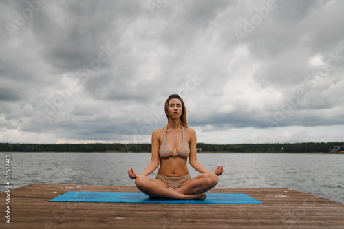 Fit slim woman practicing yoga exercises wearing mini swimsuit bikini at lake with clouds - Yoga meditation and wellness lifestyle concept - European Eastern Latvia Riga