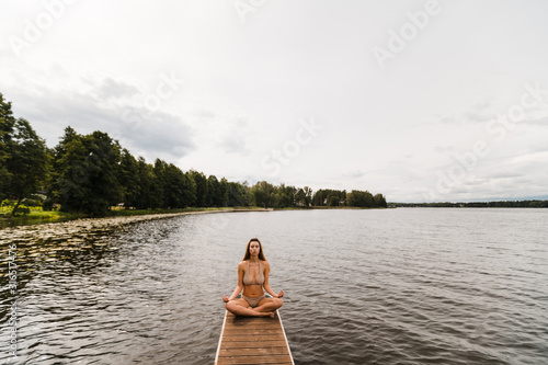 Fit slim woman practicing yoga exercises wearing mini swimsuit bikini at lake with clouds - Yoga meditation and wellness lifestyle concept - Full sitting shot