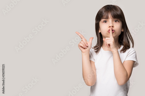 Adorable girl showing hush secret sign, pointing at copy space.