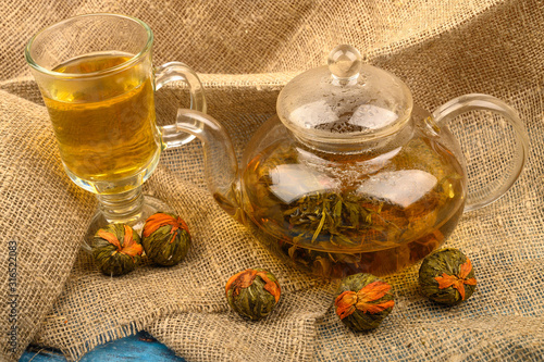 Flower tea brewed in a glass teapot, a glass of tea and balls of flower tea on a background of rough homespun fabric. Close up.