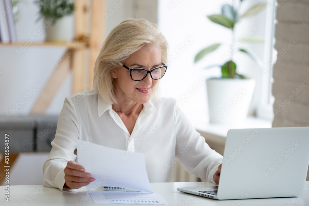 Middle-aged businesswoman typing on pc holds paper prepares report Middle-aged businesswoman typing on pc holds paper prepares report