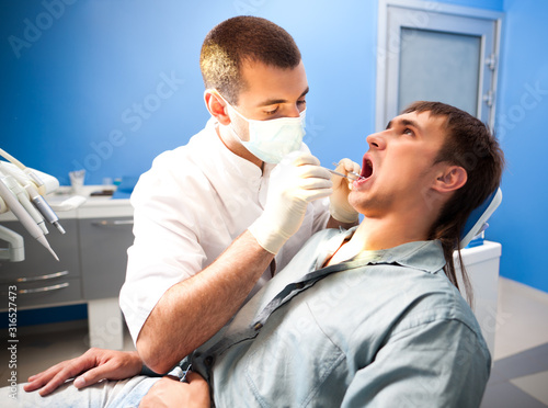 Young serious dentist doctor man in white traditional uniform and special mask sitting and examining man patient in dental office in clinic. Dental health and medical care concept